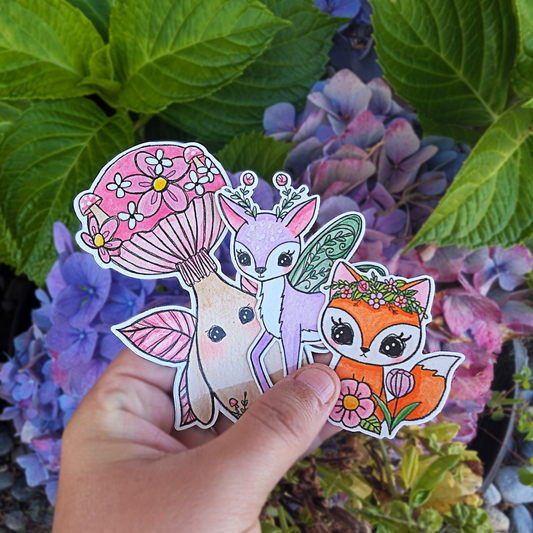 Hand holding three fairy animal stickers against a background of flowers and green leaves